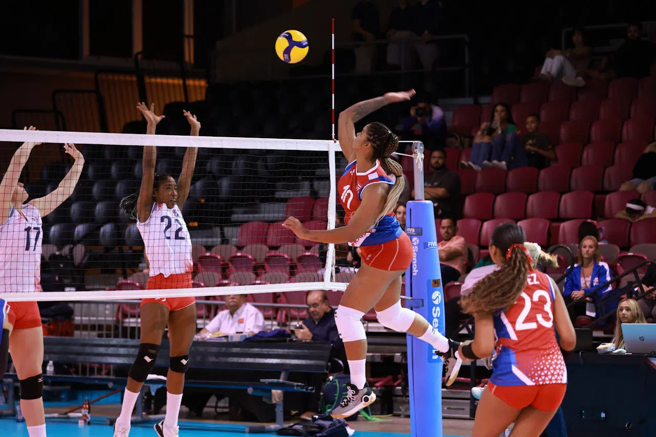 Dynamic action shot from a womens volleyball match with players in mid-air at the net.